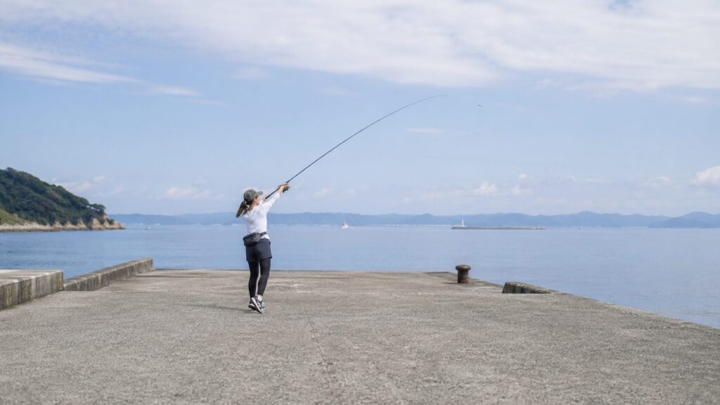 広々とした堤防の端でキャストの練習をする日本人女性の全身ショット。周囲に5メートル以上の余裕があり、釣竿を振りかぶっている動作の途中。開放的な青空と穏やかな海が背景に広がり、失敗を恐れずに練習できる安全な空間が表現されている。