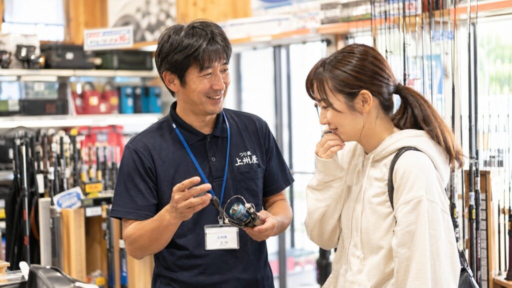 釣具屋の店内で、優しい笑顔の日本人男性店員が、緊張しながらも安心した表情の日本人女性客に釣具を説明している温かいシーン。明るい店内に整然と並ぶ釣竿やリール、タックルボックスが背景に広がる。